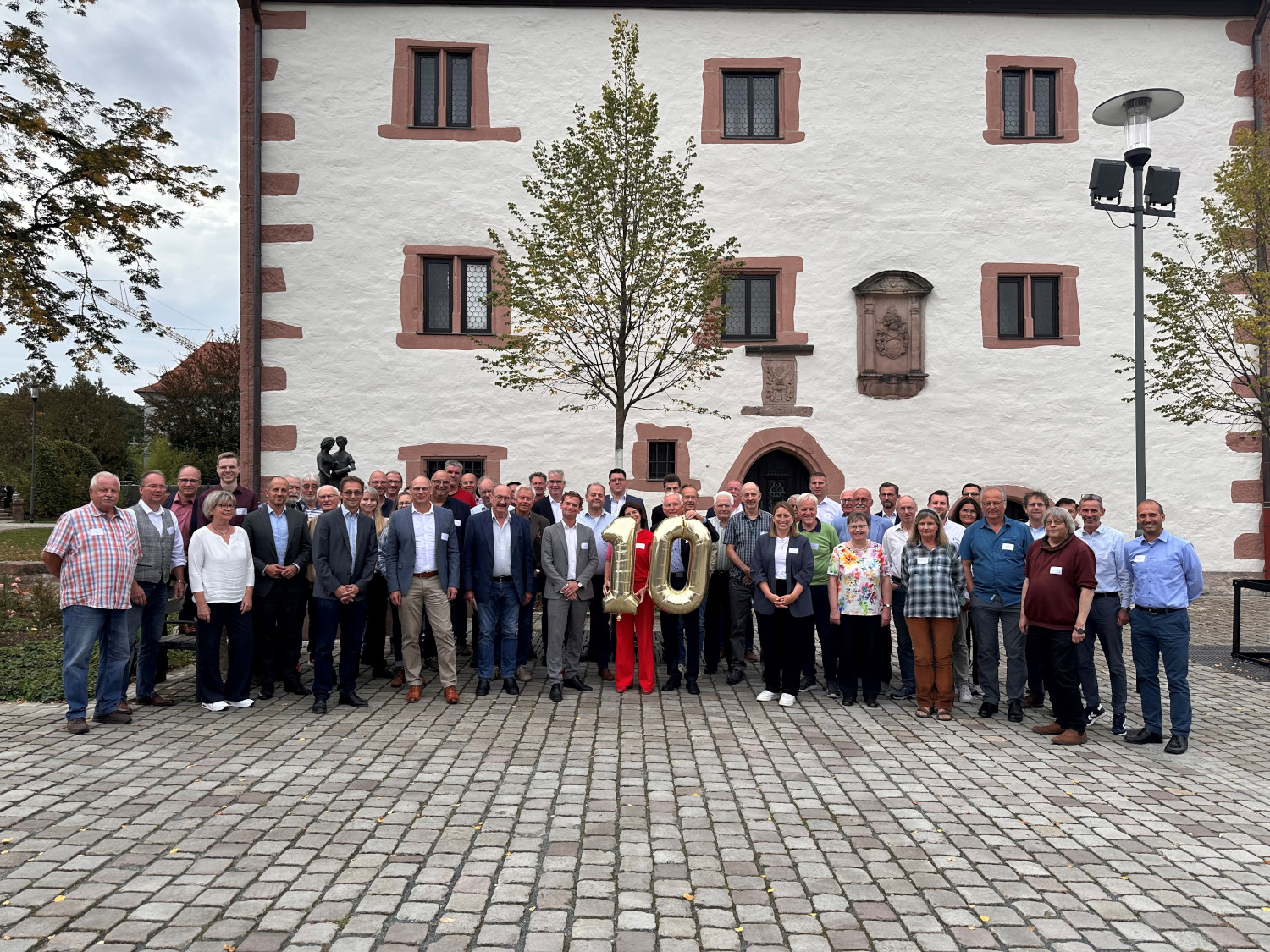 Eine große Gruppe Menschen vor einem alten Haus. Zwei Personen in der Mitte halten eine goldene 10 in Händen.